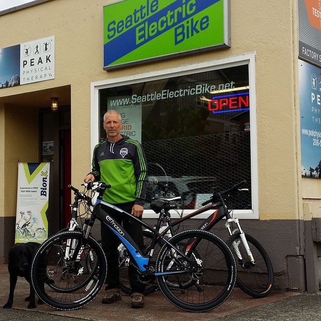 Man standing outside of the Seattle Electric Bike shop with his bike and dog.